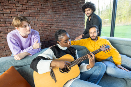 This image warmly depicts a moment of musical exchange among friends in a comfortable, homely setting. A woman is intently playing the guitar while her friends watch and listen, creating a casual and intimate atmosphere. The brick wall in the background adds a rustic charm to the scene, which is lit by natural daylight streaming through the adjacent window. Friends Sharing a Musical Moment with Guitar on a Cozy Couch. High quality photoの写真素材