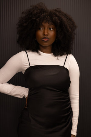 A woman with black hair wearing a black dress and white shirt is standing in front of a black wall, showcasing a trendy fashion designの写真素材