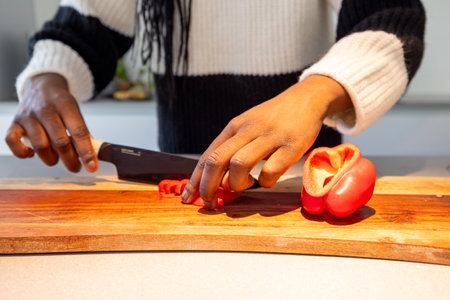This close-up image captures the hands of a Black woman as she skillfully chops a red bell pepper on a wooden cutting board. The focus on her hands highlights the culinary activity, set in a well-lit kitchen with a contemporary design, adding a warm, inviting feel to the scene. Close-up of Hands Chopping Red Bell Pepper on Wooden Board. High quality photoの写真素材