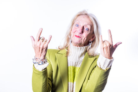 A cheerful elderly woman in a green jacket, looking joyful and relaxed, is making rock and roll hand...