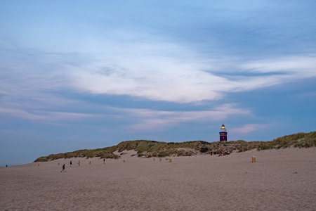 In the soft light of the early evening, several individuals take a leisurely stroll along the expansive sandy beach, with the dunes rising gently in the background. A prominent lighthouse, its vivid color diminished by the distance, stands as a silent guardian over the scene. The sky above is a tapestry of blue and white, with clouds stretched thin by the coastal breeze, suggesting a peaceful transition from day to night. Evening Stroll by the Dunes with Distant Lighthouse View. High quality photoの写真素材