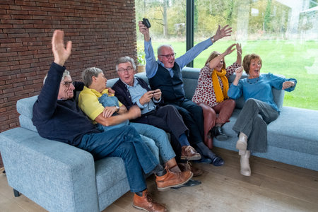 Elderly friends are happily bonding and laughing together while watching TV on a cozy sofa in a modern homeの写真素材