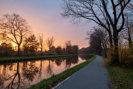 This image presents a serene riverside scene at sunset. The sky, painted in soft hues of pink and orange, reflects on the calm water, creating a symmetrical composition with the silhouetted trees. A paved path runs alongside the river, leading the viewers eye through the photograph and inviting thoughts of a peaceful evening walk. Sunset Glow along the Riverside Path. High quality photoの写真素材