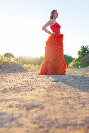 A woman in a stunning red gown walks on a sunlit rural dirt road, creating a serene and contrasting ambianceの写真素材