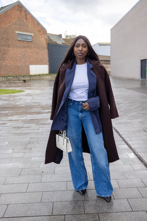 Chic street style: Woman in a brown coat, white top, blue jeans, holding a handbag on a rainy urban day.の写真素材