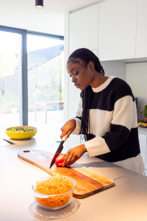 A young woman is slicing vegetables on a wooden cutting board in a modern kitchen flooded with...