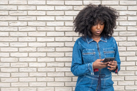Young woman with natural curly hair using her phone, standing against a light brick wall in a denim outfitの写真素材