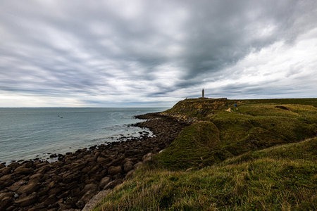 Rocky coastline with a lighthouse on a hill under a dramatic sky, offering an inspiring coastal view.の写真素材