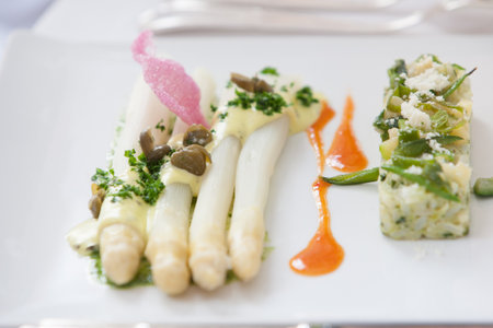 Elegant white asparagus dish with garnishes, accompanied by a side and decorative sauces on a plate.
