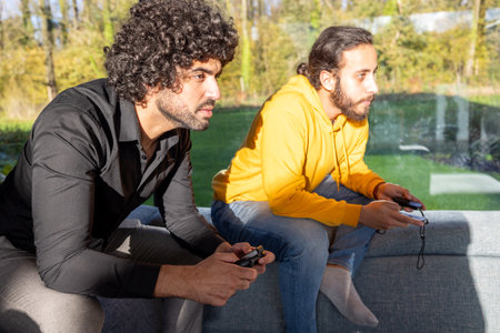 In a welllit living room, young men with focused expressions are playing video games together on a sofaの写真素材