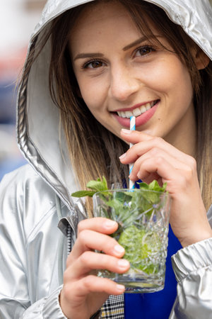 A young woman, smiling, enjoys a refreshing mint cocktail with a striped straw, wearing a street style jacket outdoorsの写真素材
