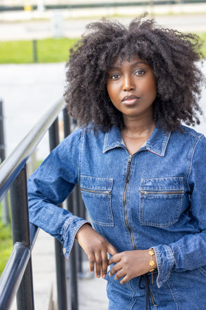A confident woman with natural curly hair is wearing a denim outfit and posing outdoors, leaning on...