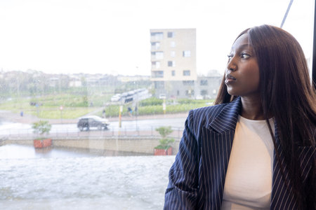 A professional woman looking thoughtfully out a window in a modern office setting with an urban landscape.の写真素材