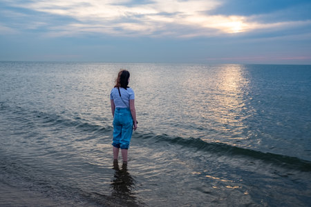 The image features a person in a moment of solitude, standing in the shallow waters of the sea as dusk settles in. The individual is dressed in rolled-up jeans and a casual shirt, suggesting a relaxed end-of-day retreat to the waters edge. The vast ocean extends into the distance, meeting the subtle gradations of twilight colors in the sky. The gentle waves around them reflect the fading sunlight, creating a path of shimmering light that leads to the setting sun partially hidden behind the clouds. Solemn Contemplation at Dusk by the Sea. High quality photoの写真素材