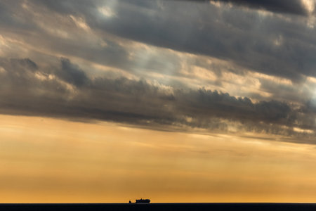 A lone ship sails under a dramatic cloudy sunset, with vibrant orange hues and dark clouds over the...