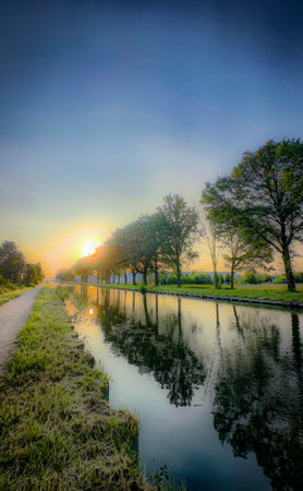 Experience a stunning sunrise over a tranquil canal, surrounded by lush trees and open fields, a picturesque sceneの写真素材
