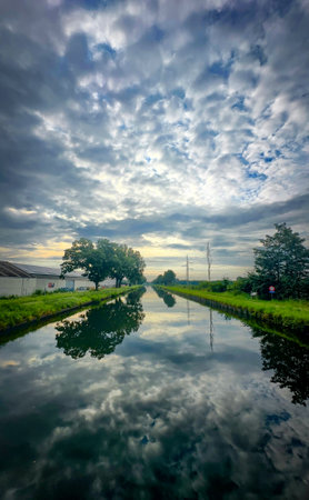 A peaceful and tranquil canal scene with beautiful reflections of clouds and trees under a dramatic sky aboveの写真素材