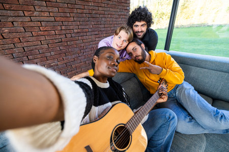 Four friends bond over music in a cozy living room, taking a selfie as one plays the guitar, creating joyful memoriesの写真素材