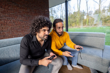 In a modern living room with natural light, two friends are deeply engaged in playing video games on the couchの写真素材