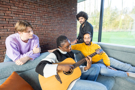 A group of friends is relaxing on a sofa in a cozy, modern living room, while one of them plays an acoustic guitarの写真素材