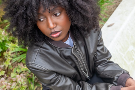 A contemplative African American woman with a full afro sits outdoors, her distant gaze and casual pose against the garden setting convey a moment of solitude and inner reflection. Pensive Reflections: African American Woman with Afro in Outdoor Solitude. High quality photoの写真素材