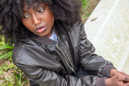 A portrait of a contemplative African American woman sitting outdoors, her expressive eyes and the natural curl of her hair highlighted against a soft, natural background. Shes dressed in a leather jacket, suggesting a blend of thoughtfulness and style. Pensive African American Woman with Natural Hair Outdoors. High quality photoの写真素材