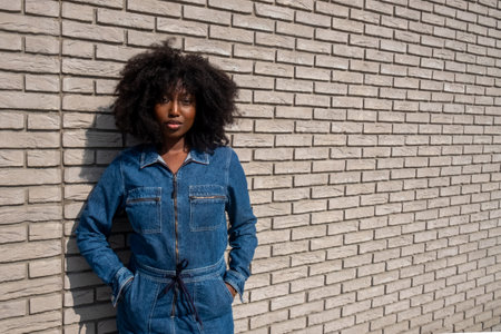 An African American woman with natural afro-textured hair stands confidently against a white brick wall backdrop. She sports a stylish full denim jumpsuit that cinches at the waist, complementing her strong stance and the urban, modern vibe of the scene. Confident Young Woman in Chic Denim Outfit Against Brick Wall. High quality photoの写真素材