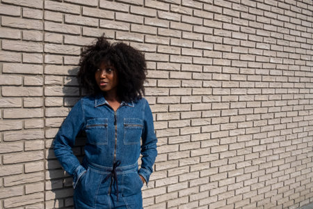 A stylish young African American woman with a voluminous afro hairstyle stands casually against a textured brick wall, her hands tucked into the pockets of her chic denim jumpsuit. Her sideways glance and relaxed demeanor evoke an air of effortless urban cool. Young African American Woman Posing in Trendy Denim Outfit. High quality photoの写真素材