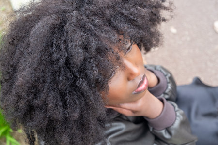 An intimate perspective of a young African American woman looking upwards, her voluminous afro framing her dreamy expression. A sense of wonder and daydreaming is captured against a blurred urban backdrop. Dreamy Gaze: Upward Looking African American Woman with Voluminous Afro. High quality photoの写真素材