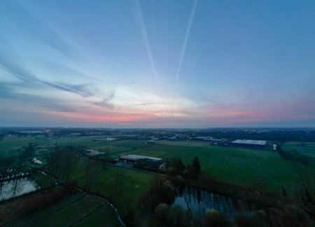 An aerial view captures the sprawling industrial greenhouses as day yields to night. The sunset casts a warm glow over the landscape, creating a striking balance between the industrial elements and the natural serenity of the countryside. High quality photoの写真素材