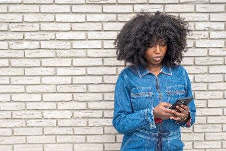 A young African American woman stands focused on her smartphone against a white brick wall backdrop. Her curly afro frames her face while her denim outfit reflects a casual, urban style, depicting a moment of modern daily life. Young African American Woman Texting on Smartphone Against White Brick Wall. High quality photoの写真素材
