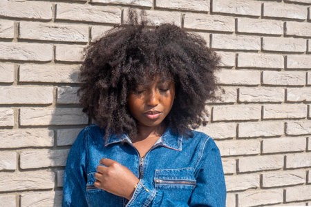 Captured in contemplation, a young African American woman with a full, natural afro stands against a stark white brick wall. The sunlight highlights her denim jacket, adding warmth to the image, while her thoughtful expression suggests an introspective moment. Pensive Young African American Woman in Denim Attire Against White Brick Wall. High quality photoの写真素材