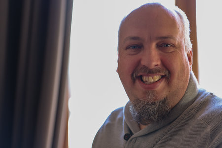 A cheerful man with a distinct beard smiles warmly in a bright, welllit room by a large window nearbyの写真素材