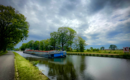 A charming canal scene with a slim barge navigating lush greenery and dramatic clouds, creating an...