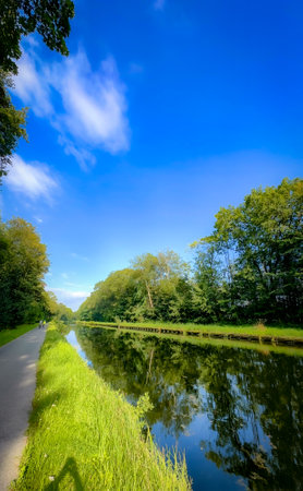 A serene and tranquil canal scene showcases clear reflections, lush green banks, and radiant blue skies aboveの写真素材