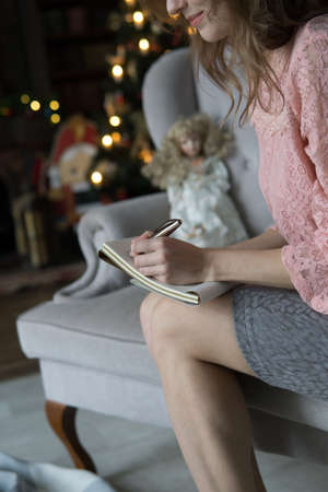 young blonde woman sits on a gray sofa in a pink blouse and writes with her left hand with a pen in...