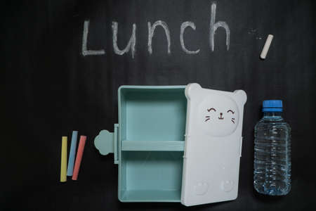 open an empty lunch box with a sweet little face and a bottle of water on a black background with colorful crayons and lunch inscription written in white chalk.の写真素材