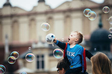 KRAKOW, POLAND - JUNE 27, 2015: Adorable little boy sits on dad's shoulders and touches the giant bubbles at the street.のeditorial素材