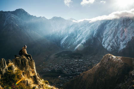 Young tourist in bright hat, black trousers with a backpack sit on cliff's edge and looking at the misty mountain village and glacier at sunrise, Stepantsminda, Georgiaの写真素材