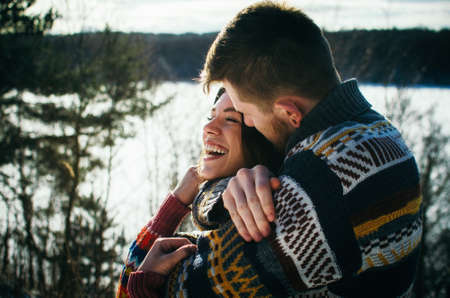 Joyful cute couple embraces. Young man in a sweater hugs a girl from behind background in winter. The concept of a successful relationship and happy momentsの写真素材