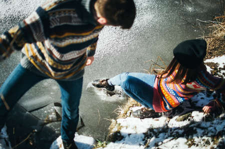 Young woman using a shoe breaks the ice while standing on the shore with boyfriend. Winter and...
