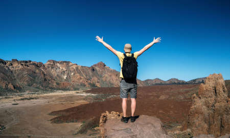 Man with a backpack stands back to the camera with raised hands. Traveler are enjoying the landscape with mountain valley of the crater of Teide volcano, Tenerife, Canary islands, Spainの写真素材