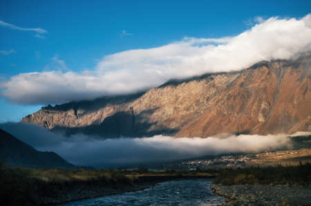 Different double layers of clouds over Stepantsminda Kazbegi village and peak of brown mountain, Georgia. Scenic autumn landscape.の写真素材