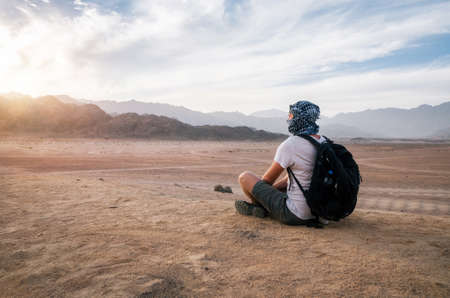 Traveler wearing arabic head scarf with a backpack sits and looks at Sinai desert and mountains at sunset, Egyptの写真素材