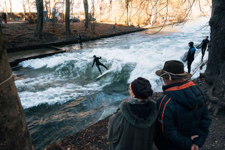 Munich, Germany - December 7, 2017: Elderly couple watch as surf ride artificial wave on the Eisbach, dangerous water Isar river channel at Englischer Gardenのeditorial素材