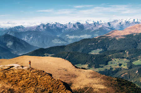 Young man hiker in bright jacket stands on cliff at mountains Dolomites and takes photography of valley, Italy, Seceda. Traveler enjoys landscapeの写真素材