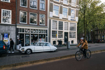 Amsterdam, Netherlands - 25 April, 2017: Adult woman rides a bicycle against parked vintage Citroen DS in historical part of Amsterdam with typical traditional houses.のeditorial素材