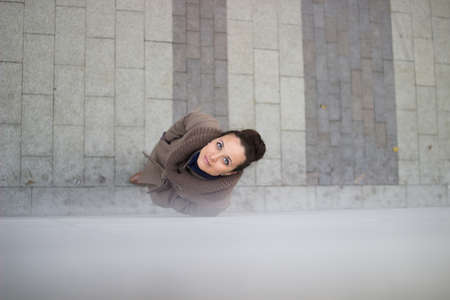 Young woman in a beige coat, stopped near the wall to look up, looks at the camera, she smiles