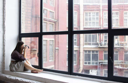 a young woman sits on a windowsill with large windows and takes picturesの写真素材
