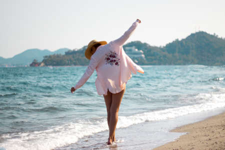 a young girl in a pale pink shirt and a straw hat walking along the seashore. Holidays. Recreation. The sun. Ocean. Sea. Shooting from the backの写真素材